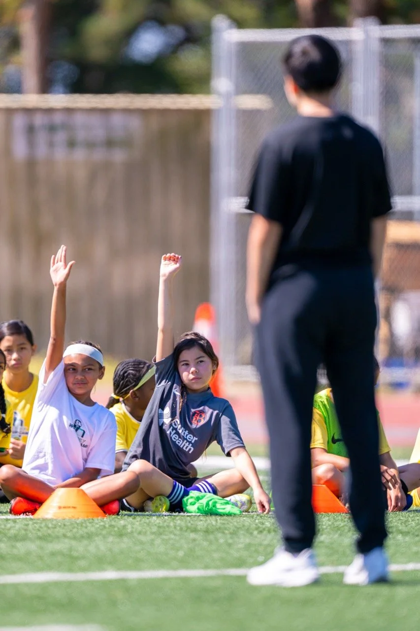 Young girls sitting on a sports field, some raising their hands, listening to a coach or instructor during a sports practice or training session.