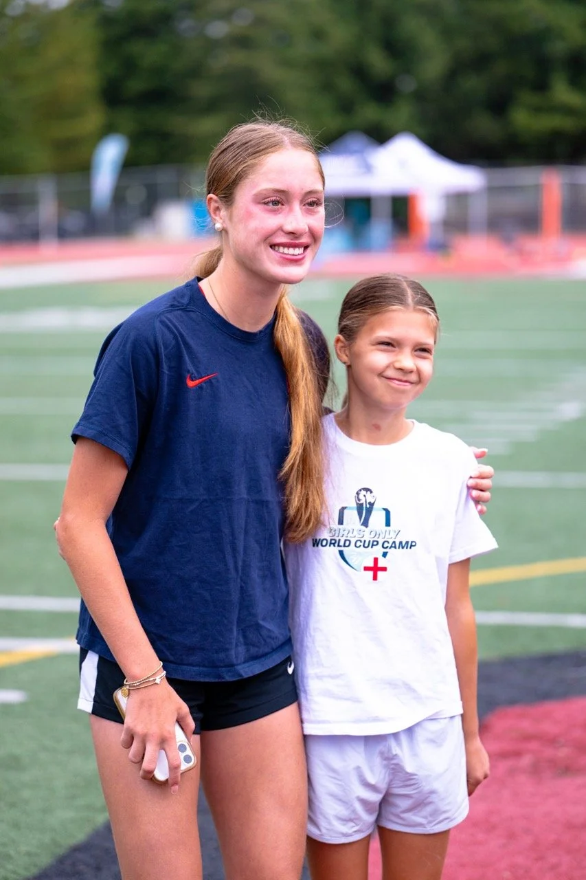 Two girls standing on a sports field, smiling, with trees and tents in the background.