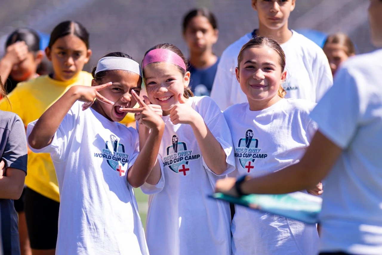 Group of young girls at a soccer camp, wearing white shirts with a logo that says "Girls Only World Cup Camp," smiling and posing for a photo on a sunny day.