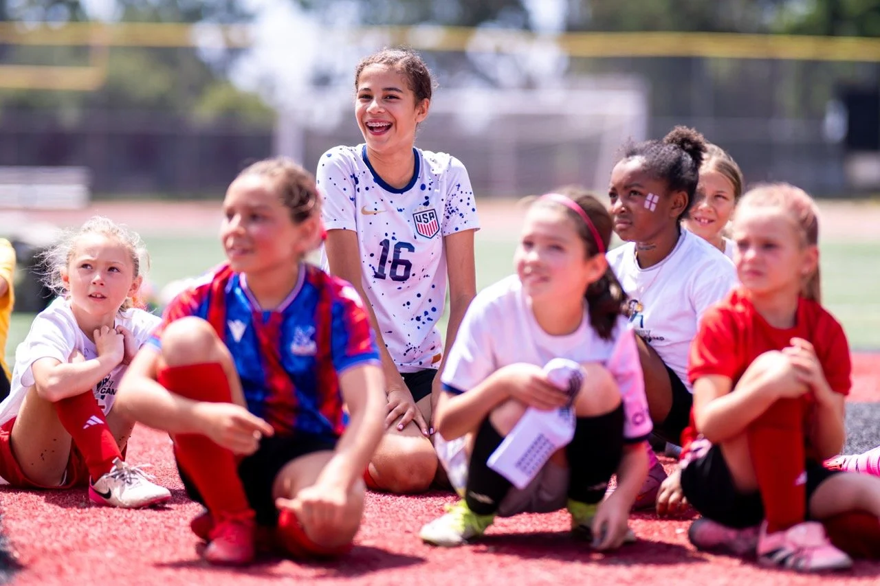 Young girls, including one in a soccer jersey with the USA logo, sitting on a sports field during a soccer practice or game.