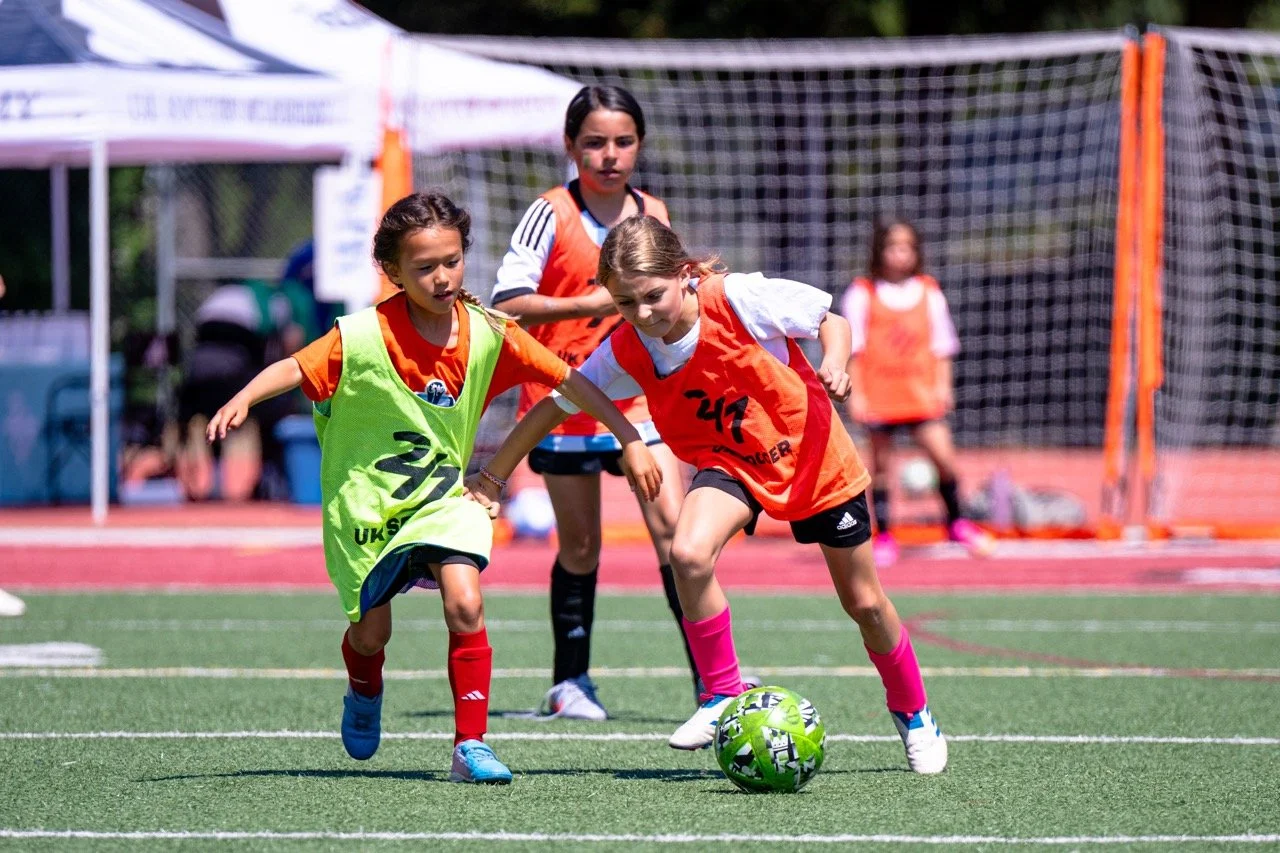 Young girls playing soccer on a field during daytime, with one girl dribbling the ball while others run behind her.