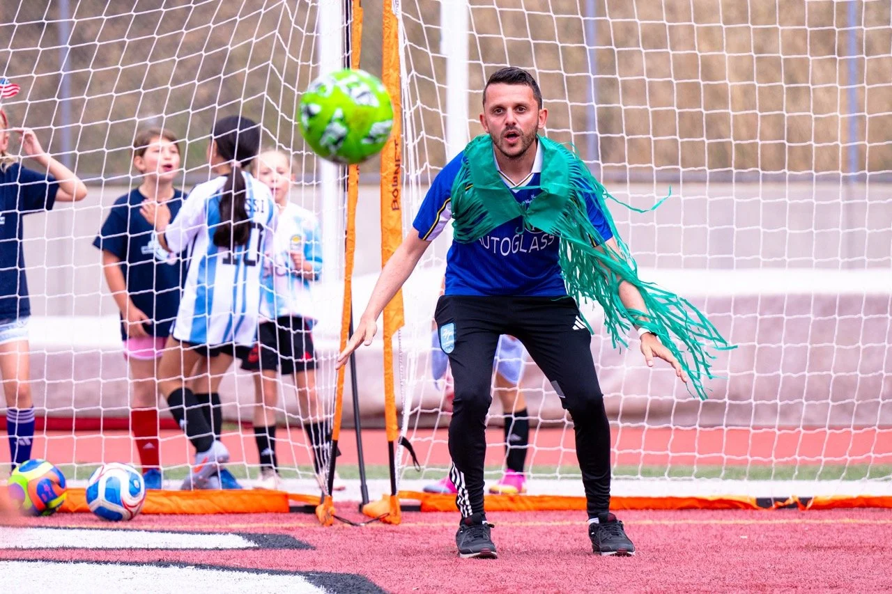 A man in a blue sports jersey and black pants playing soccer on a field, with children in jerseys behind a goal net watching in surprise.