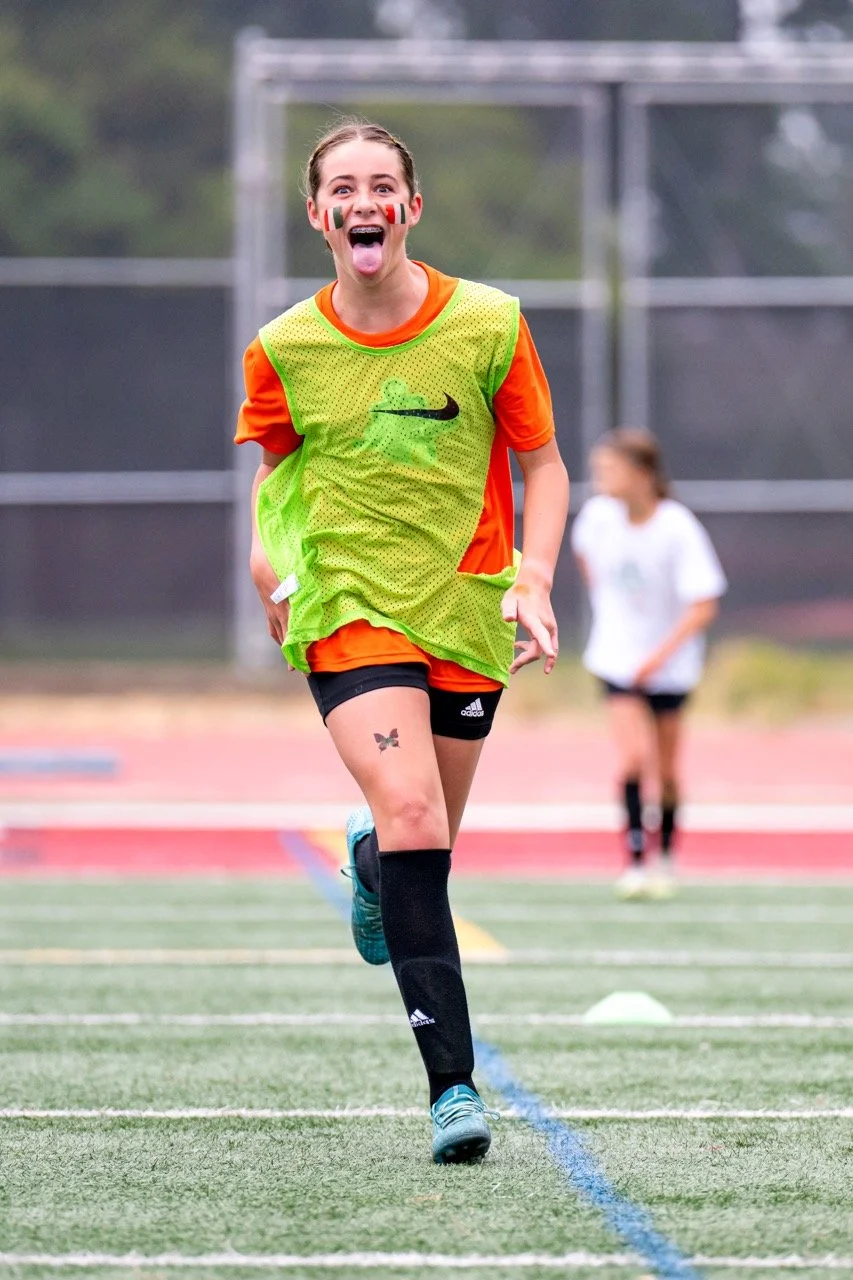 A young female soccer player with face paint, sticking her tongue out, running on a soccer field in a green and orange jersey, black shorts, and blue cleats, with another player in white in the background.