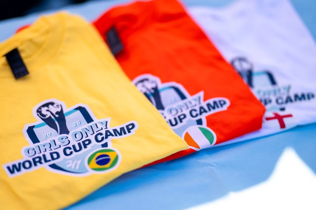 Three colorful girls' soccer jerseys in yellow, orange, and white with Girls Only World Cup Camp logos on a blue surface.
