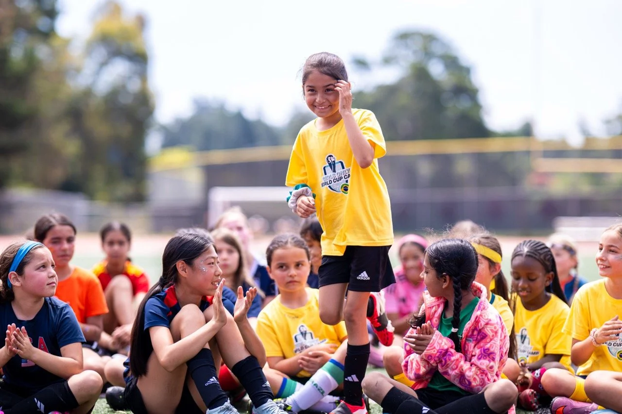 A group of young girls sitting on a sports field, with one girl in a yellow shirt standing up and smiling while others watch and clap.