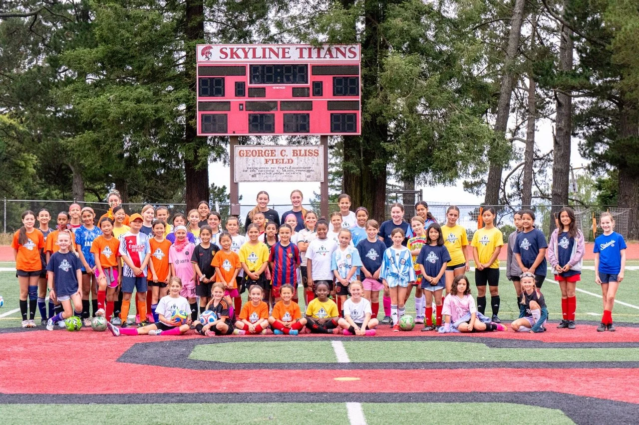 Group of young girls and coaches on a soccer field posing for a team photo with a scoreboard and trees in the background.