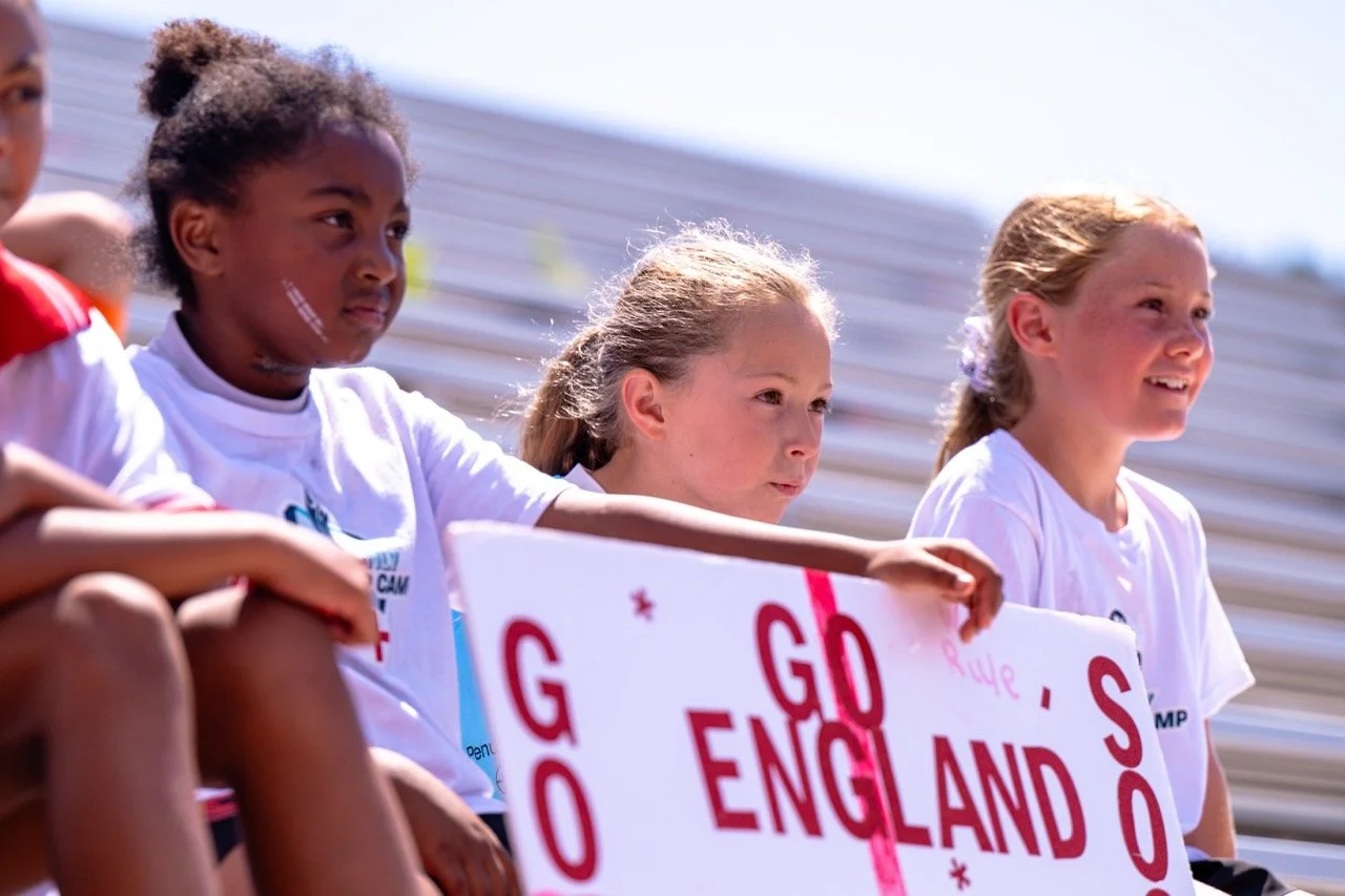 Young girls sitting on bleachers at a sports event, holding a sign that says "Go England"; they are wearing white t-shirts and have focused and happy expressions.