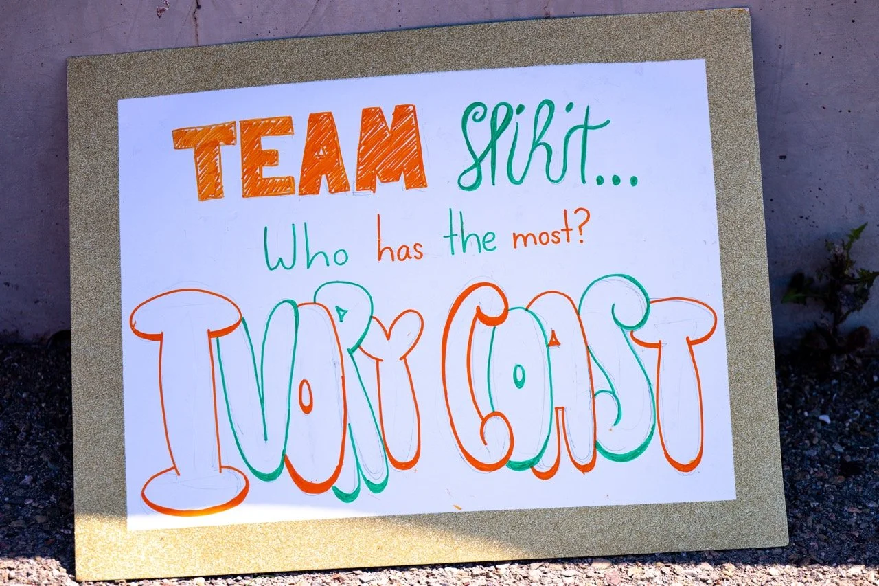Handwritten sign on a corkboard asking 'Who has the most?' about a team shirt, with colorful letters spelling 'I LOVE COAST'.
