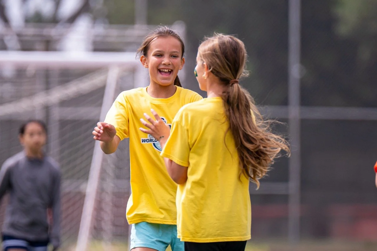 Two young girls in yellow shirts sharing a happy moment on a soccer field, surrounded by a fence and soccer goal, with a third girl in the background.