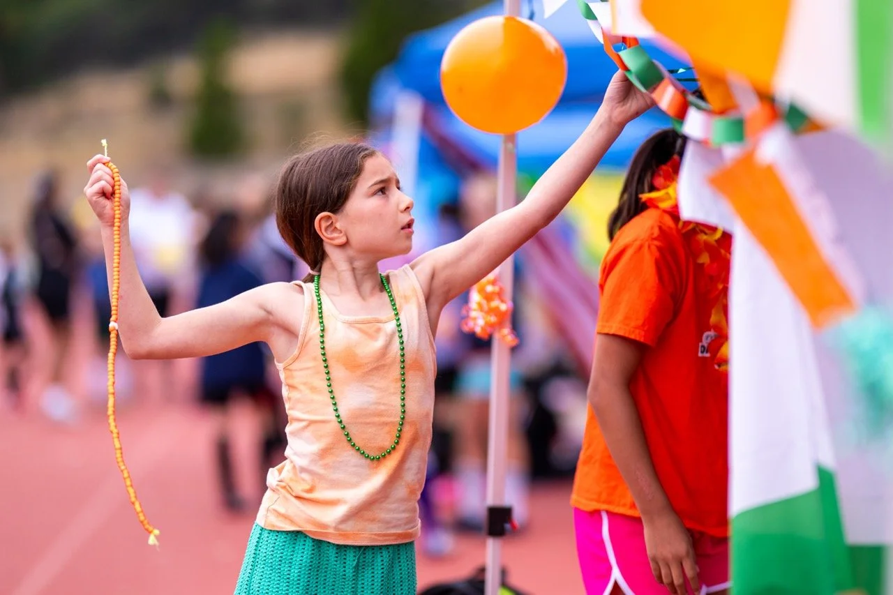 A young girl at an outdoor event holding a string of beads in one hand and reaching toward a bright orange prize balloon with the other, surrounded by colorful decorations and other people in the background.