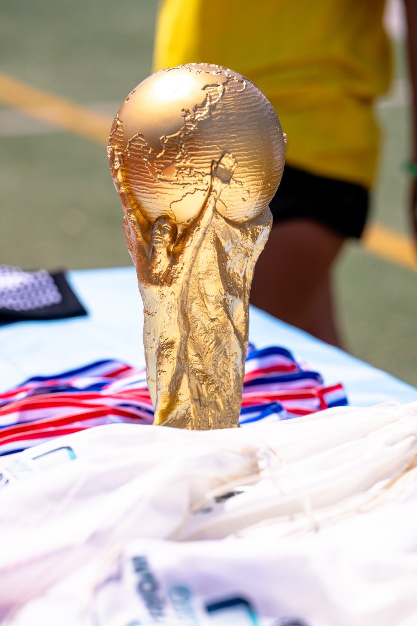 Close-up of a gold FIFA World Cup trophy with a figure holding up a globe, placed on a table with medals in the background, at an outdoor sports event.