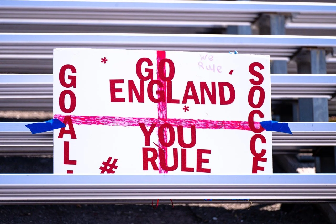A protest sign with red text on a white background reads 'GO ENGLAND SOCCER' and 'GOAL! YOU RULE'. Pink crosses and handwritten notes are also visible on the sign, which is taped to metal bleachers.
