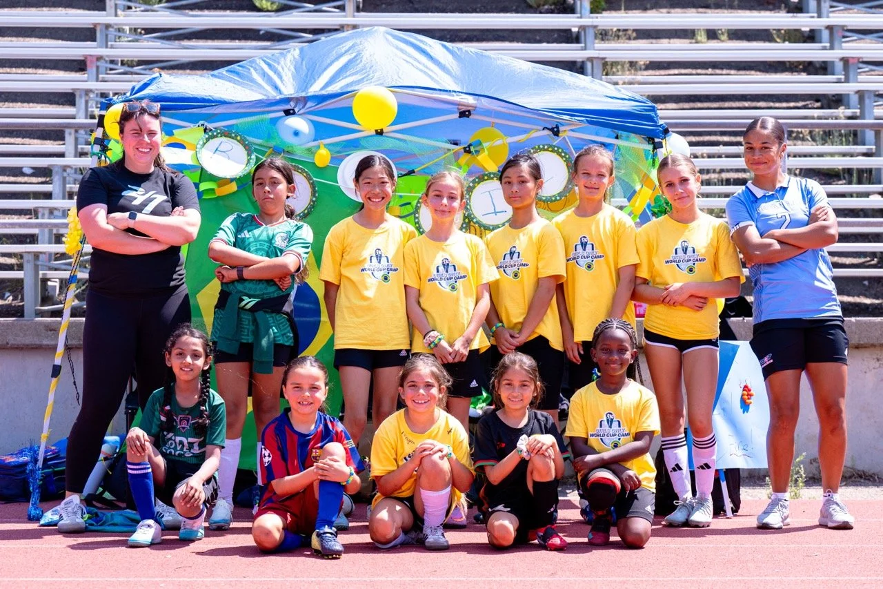 Group of young girls and two coaches in sports uniforms posing in front of a colorful decorated backdrop at a track and field stadium.