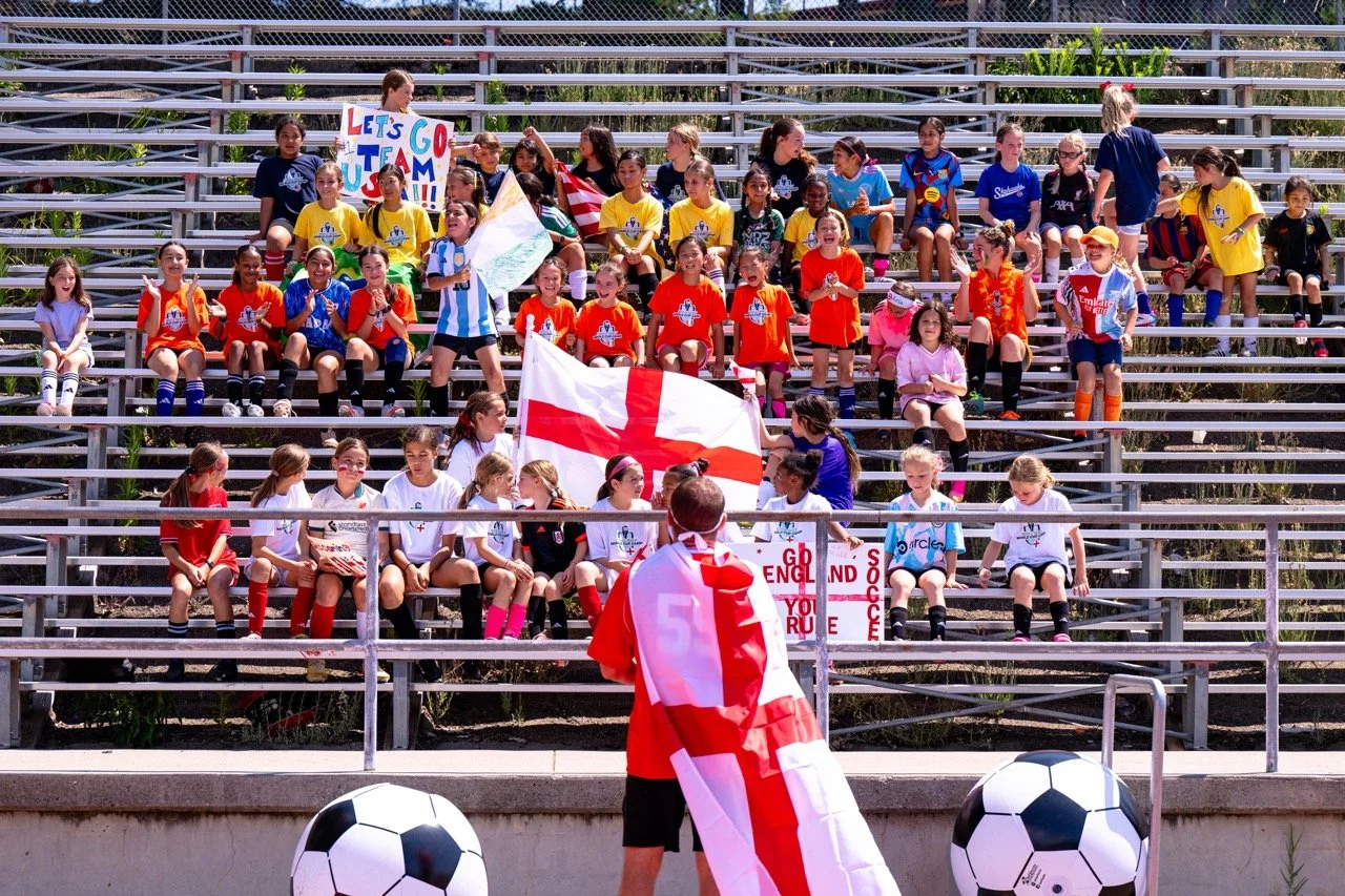 A group of young girls in soccer uniforms seated on bleachers at a stadium, holding flags and signs to support England's soccer team. A person in front draped in an England flag is addressing the group. Soccer balls are visible at the bottom of the i