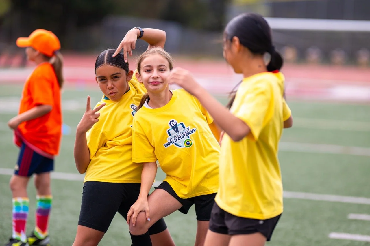 Four young girls in yellow and orange sports shirts on a field, one making a peace sign, another flexing her arm, and two talking, during a sports event or camp.