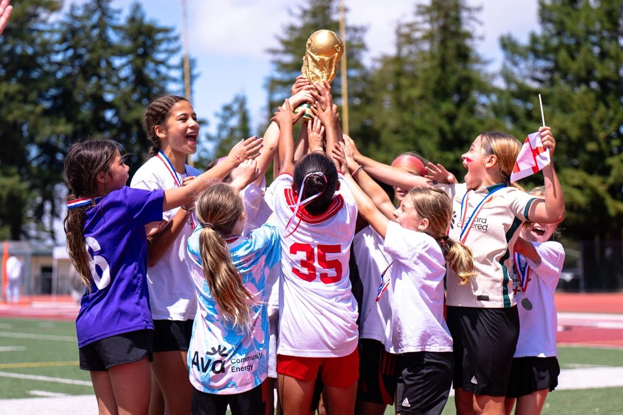 A group of young girls celebrating and lifting a golden soccer trophy on a field, wearing sports uniforms and medals.