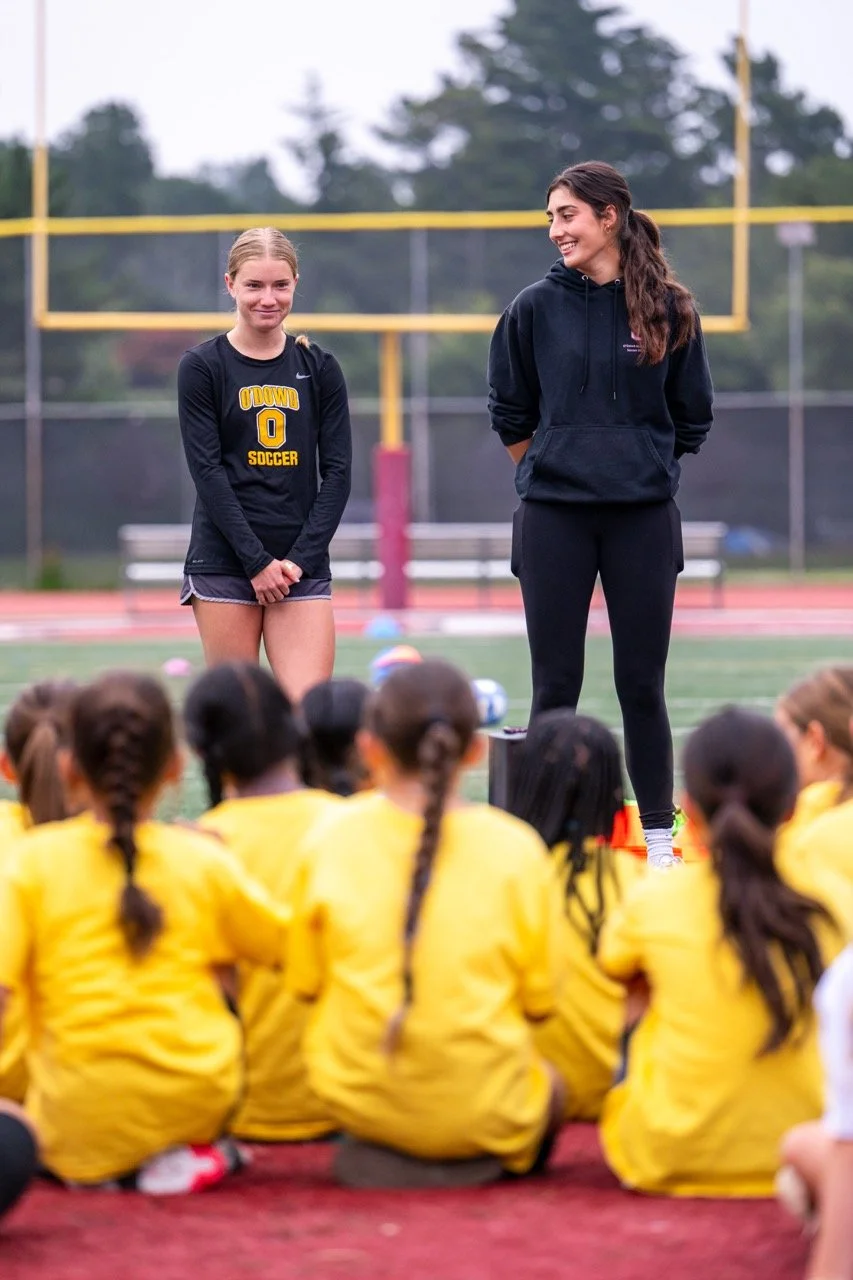 A coach speaking to a group of girls in yellow jackets on a sports field, with two girls standing nearby.