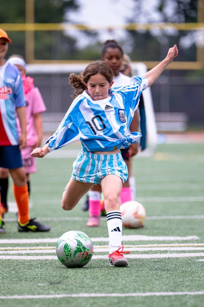 Young girl in Argentina soccer uniform kicking a soccer ball on the field, with other children watching.