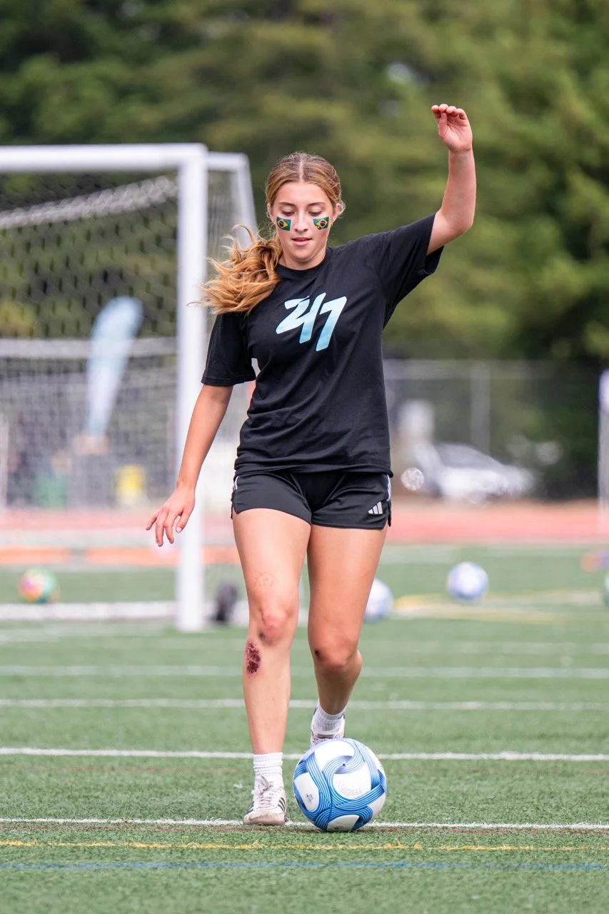 A young woman with face paint in the Brazilian flag colors, wearing a black sports shirt and shorts, is playing soccer on a field. She is about to kick a blue and white soccer ball, with a bruise and scrape visible on her left leg.