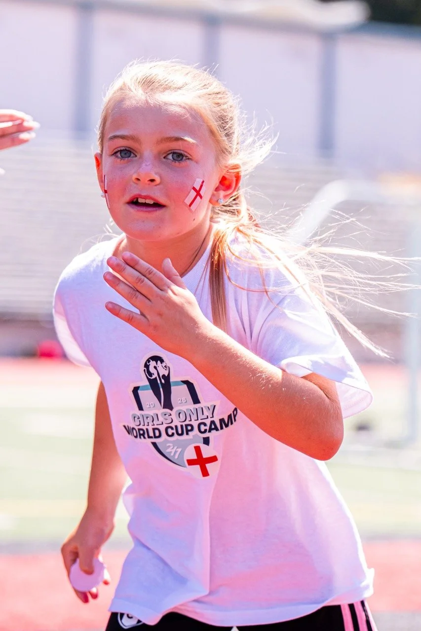 Young girl with blonde hair, face painted with an England flag, wearing a white t-shirt with a Girls Only World Cup Camp logo and a red cross symbol, appears to be participating in an outdoor event.