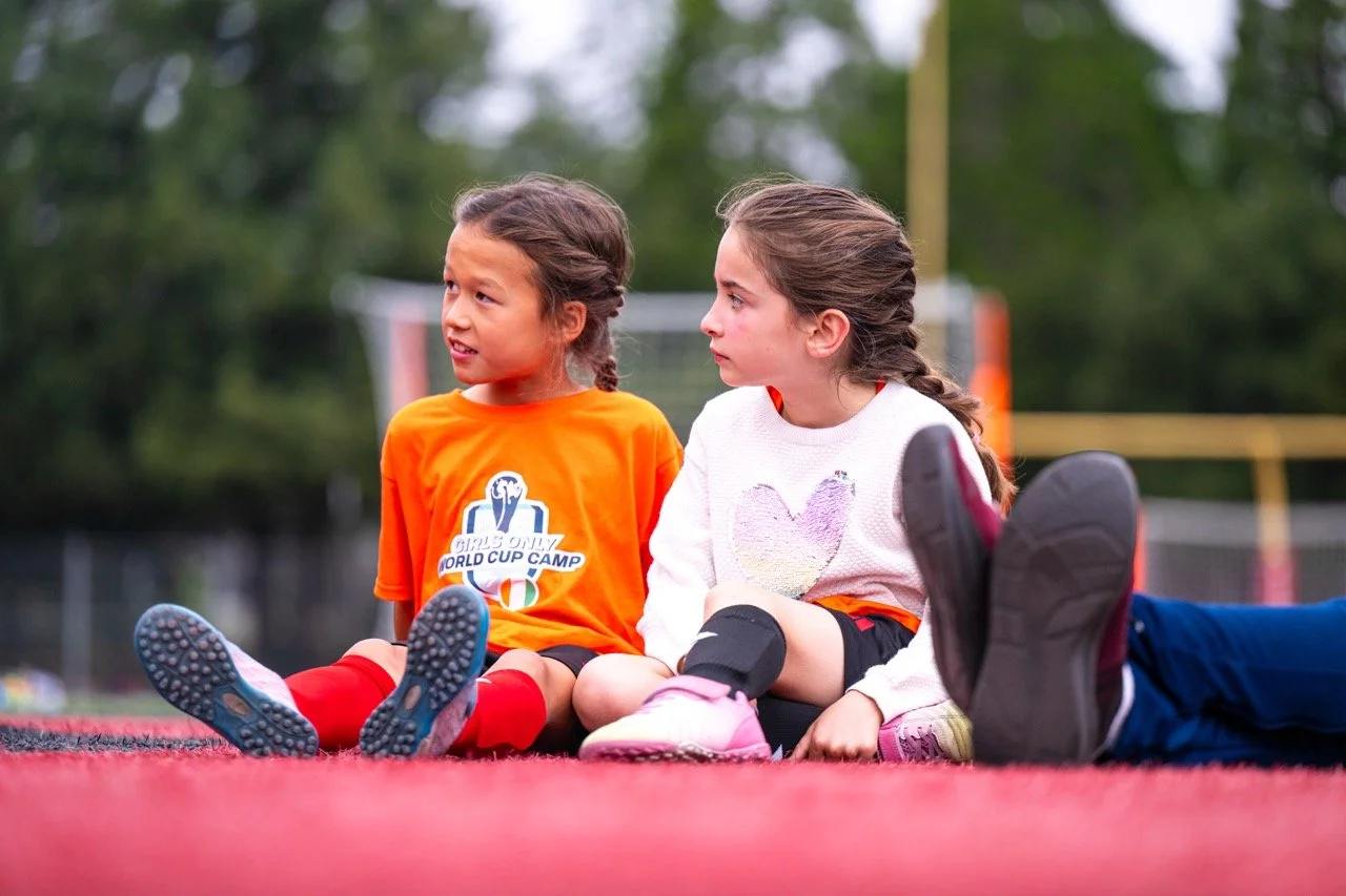 Two young girls sitting on a sports field during daytime, engaging in a conversation, with blurred trees and a sports goal in the background.