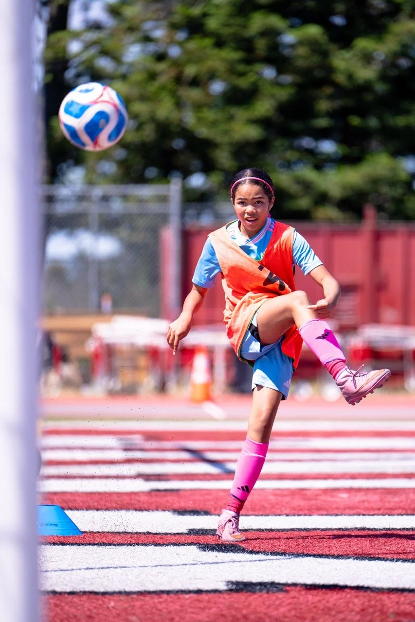 Young girl playing soccer on a field, kicking a colorful ball, wearing pink socks and sports gear, with trees and a fence in the background.
