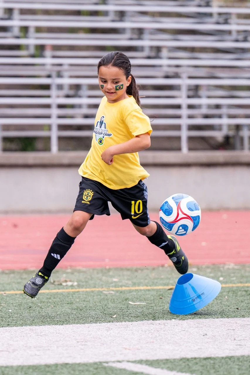 A young girl with face paint of the Brazilian flag playing soccer on a field, kicking a ball with her right foot, wearing a yellow shirt, black shorts, and black socks.