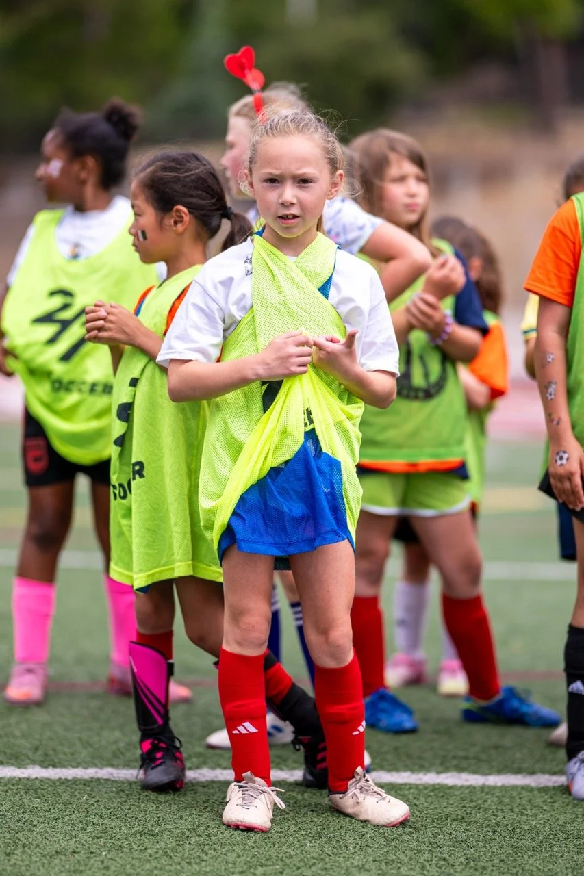 Young girl standing with other children on a soccer field, wearing a yellow training vest, a white T-shirt, blue shorts, red socks, and white shoes, with a serious expression.