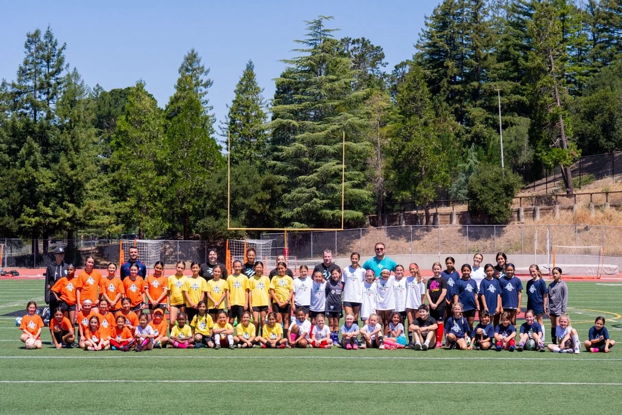 Group of young girls and their coaches on a soccer field with trees and a goalpost in the background.