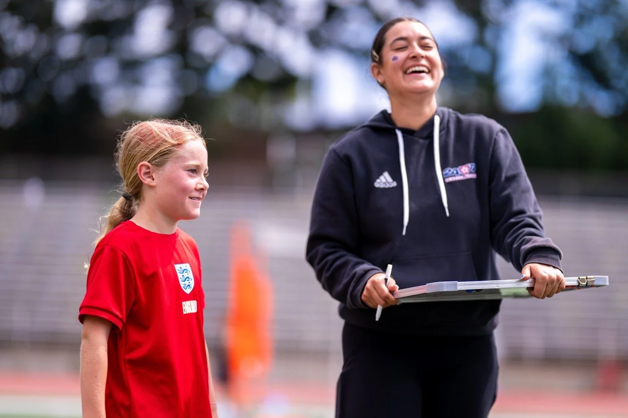 A young girl in a red sports jersey standing near a smiling coach on a sports field.