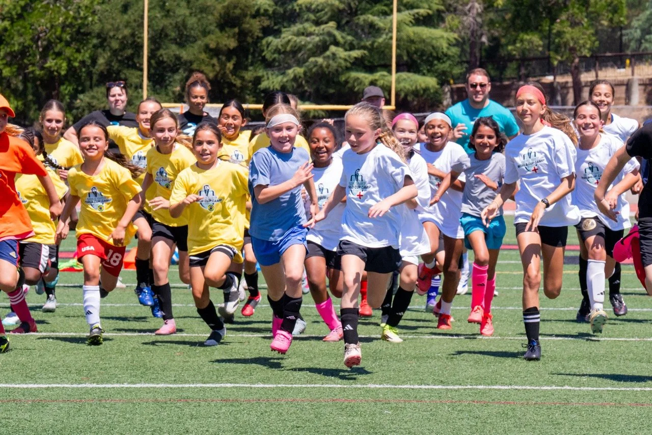 Girls running on a soccer field during a game or practice with coaches in background.