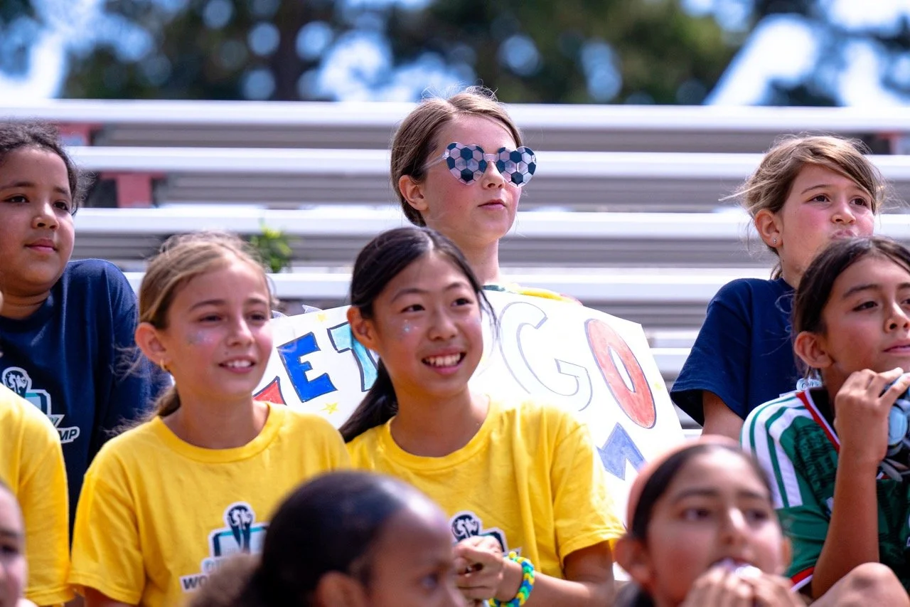 Young girls sitting in bleachers at an outdoor event, some wearing yellow T-shirts, one girl with sunglasses, holding a sign, smiling and looking happy.