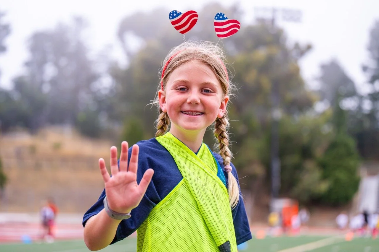 A young girl with braided blonde hair smiling and waving at the camera. She is wearing a yellow sports vest and a navy blue shirt, standing outdoors on a sports field with trees and other children in the background.