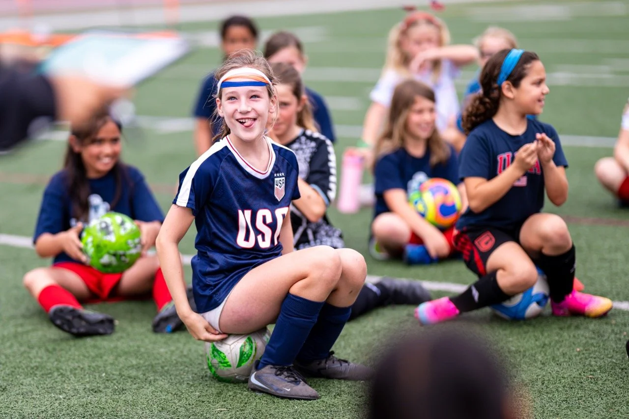 Group of young girls sitting on a sports field, wearing USA soccer jerseys, with some holding soccer balls, smiling and looking happy.
