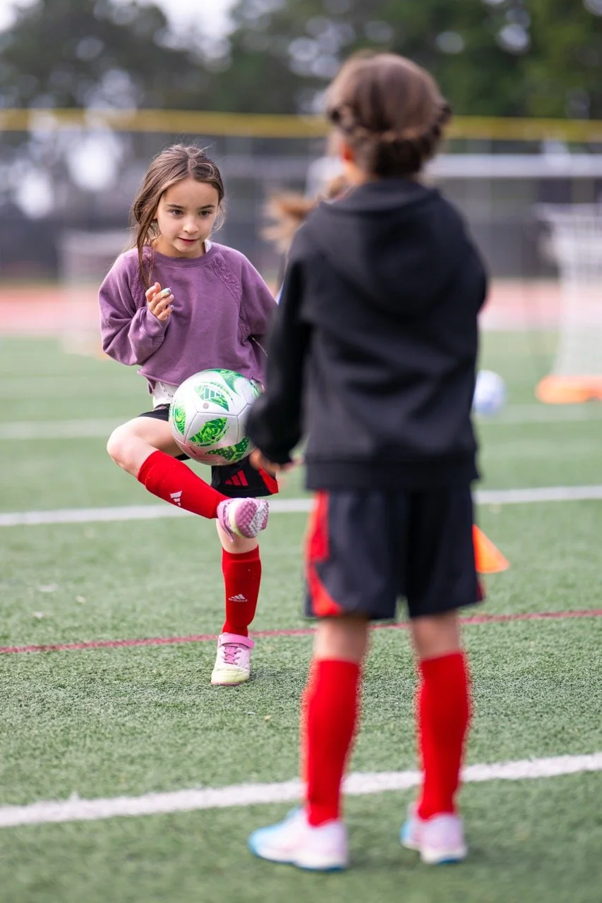 Two young girls practicing soccer on a field, one girl in a purple sweatshirt handling a ball with her foot, the other girl in a black jacket with red shorts facing her.