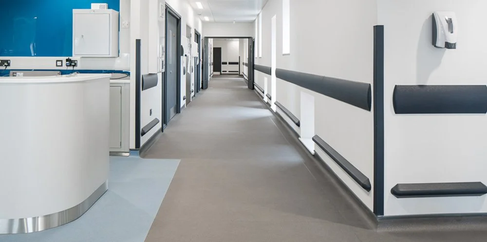 Empty hospital hallway with white walls, black wall protection handrails, and a reception desk on the left.