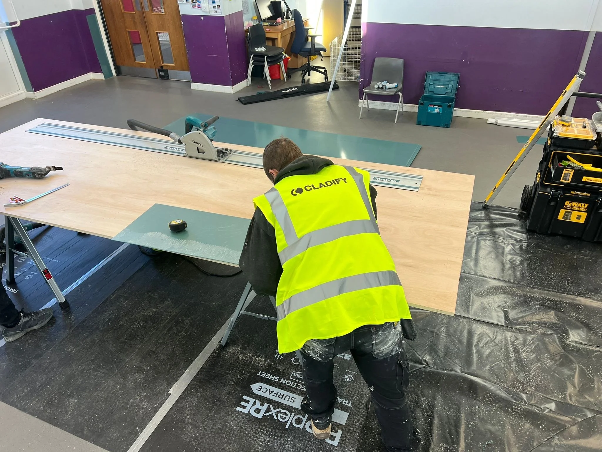 Two cladding installers workers installing hygienic cladding in a Kent-based school. One worker is kneeling and the other is standing, both wearing high-visibility safety jackets.