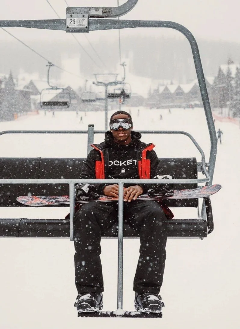 Person sitting on a ski lift chair holding a snowboard with snow falling, wearing a black jacket with red accents, black ski pants, and ski goggles, with snowy ski slopes and ski lift chairs in the background.