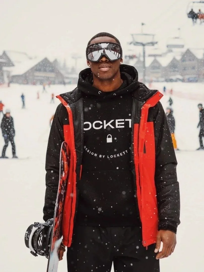 Young man standing on snow-covered ground at a ski resort, wearing ski goggles, black hoodie with 'Lockett' logo, black pants, red and black jacket, holding a snowboard, with chairlifts and other people in the background.