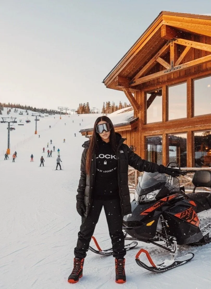 A woman in black 'Lockett' logo hoodie in ski gear standing next to a snowmobile on a snowy slope with skiers and a wooden chalet in the background.