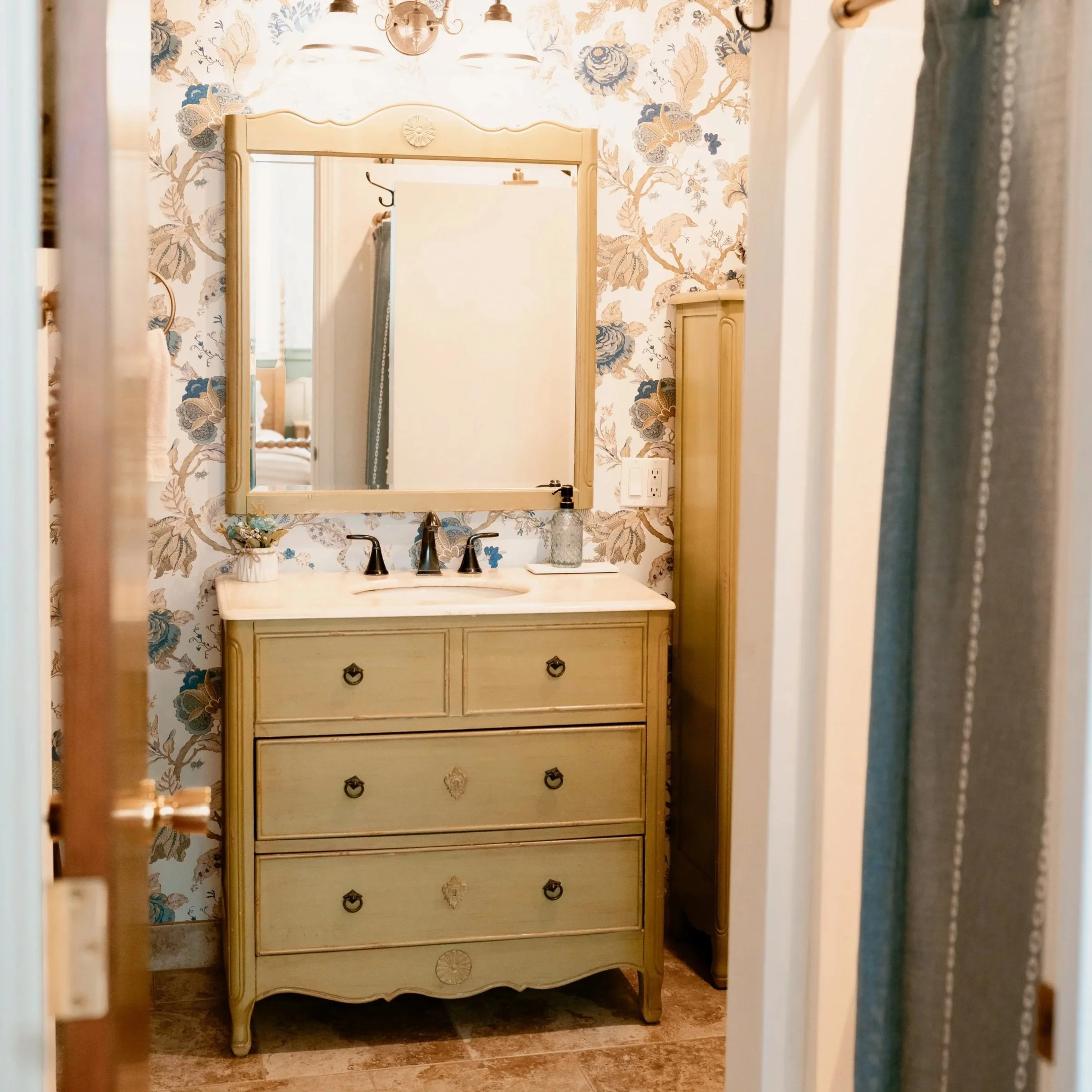 A beige vintage dresser with black handles, positioned against a floral wallpapered wall, reflecting in a mirror above a white marble countertop with a gold faucet. Decorative items include a small plant, a clear glass container, and a tray with a soap dispenser.
