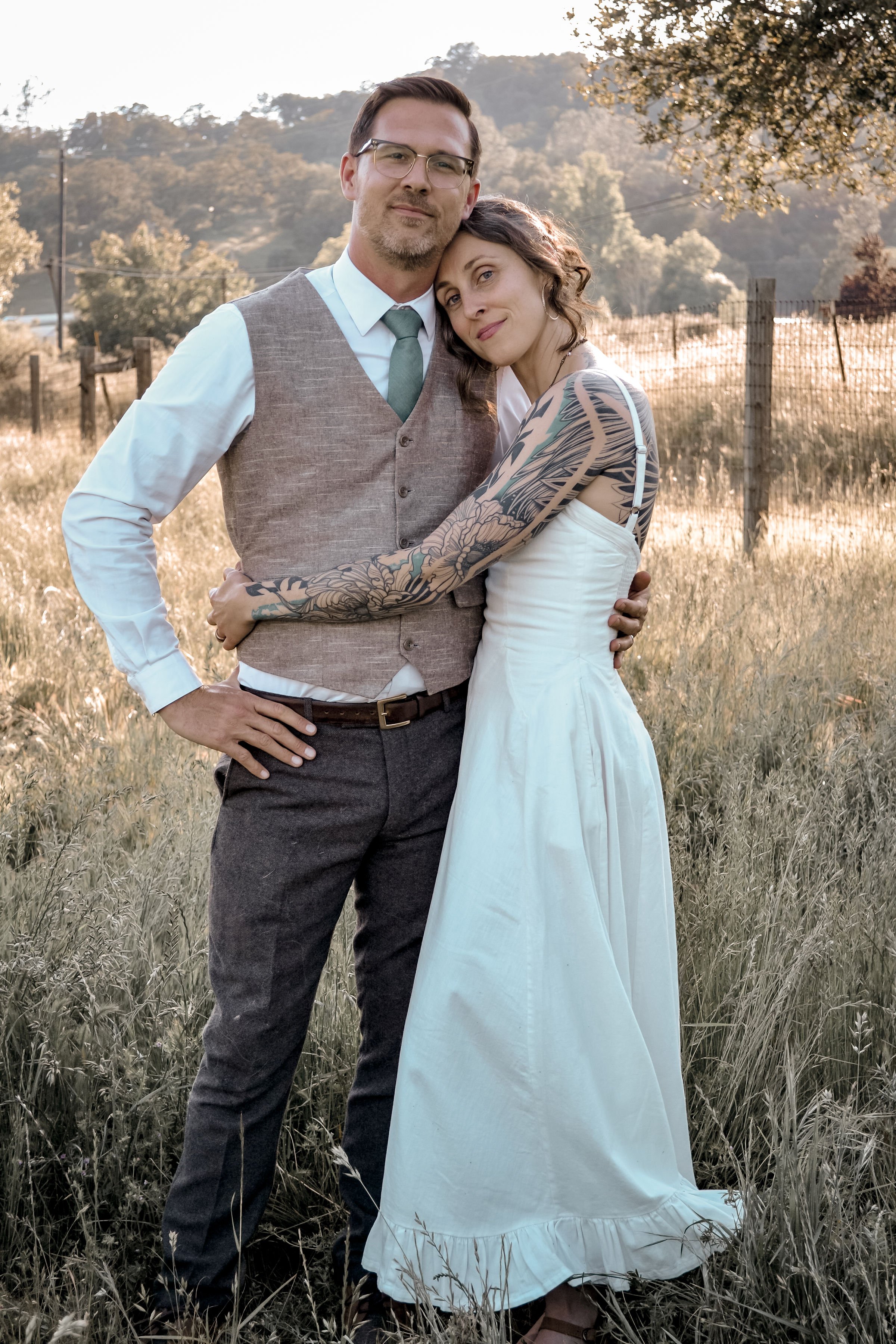 A man and woman standing together outdoors in a grassy field with trees and hills in the background during late afternoon. The man is wearing glasses, a vest, shirt, and tie, while the woman is dressed in a white dress with tattoos on her arms, leani
