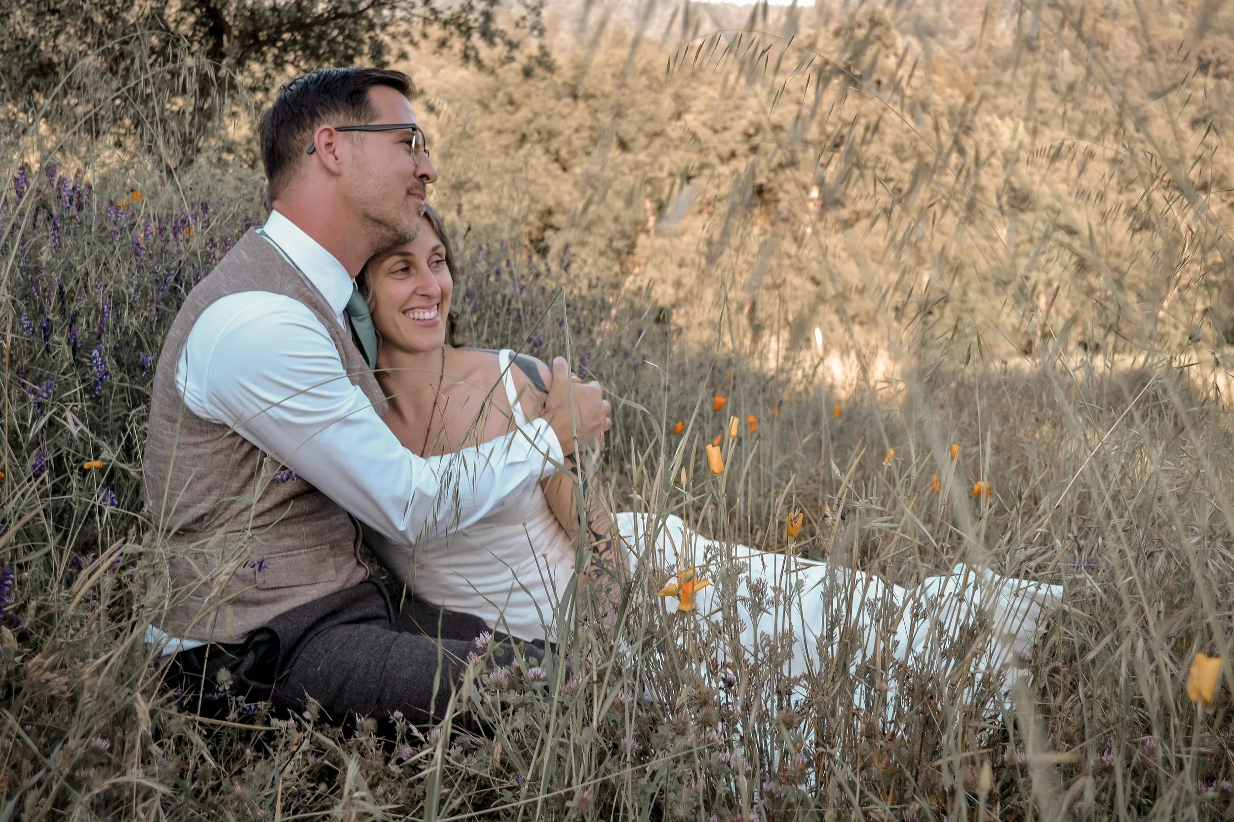 A man and woman sitting in a field of wildflowers, embracing and smiling.