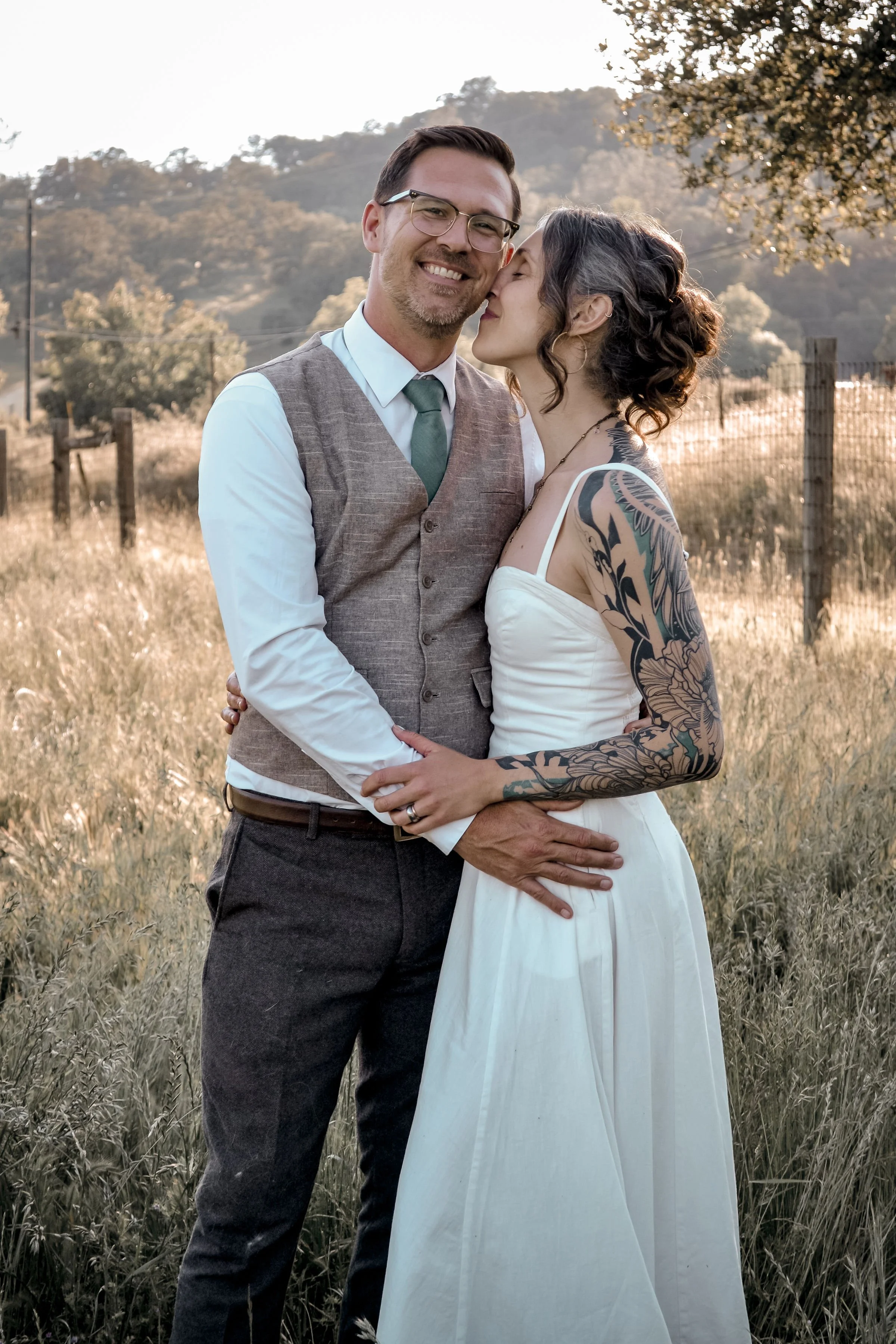 A happy couple, a man wearing glasses, a vest, dress shirt, and tie, and a woman with tattoos in a white dress, embrace in an outdoor field with mountains and trees in the background.
