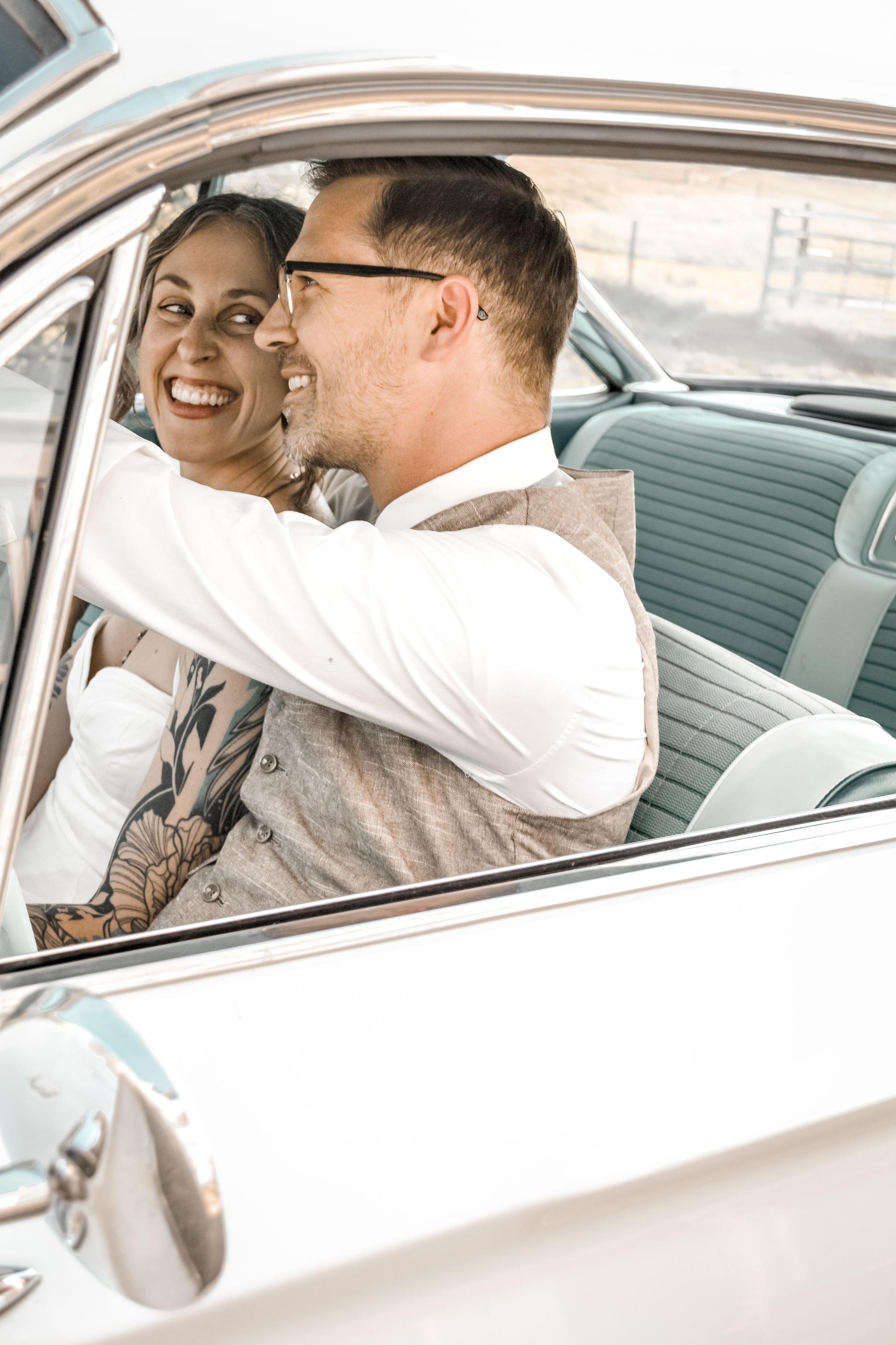 A smiling woman and man are sitting inside a vintage car, sharing a joyful moment.