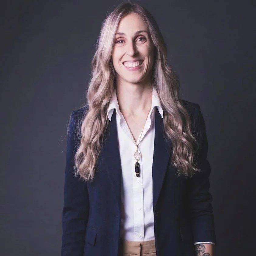 A woman with long wavy blonde hair smiling wearing a navy blazer over a white button-up shirt and a necklace, standing against a dark gray background.