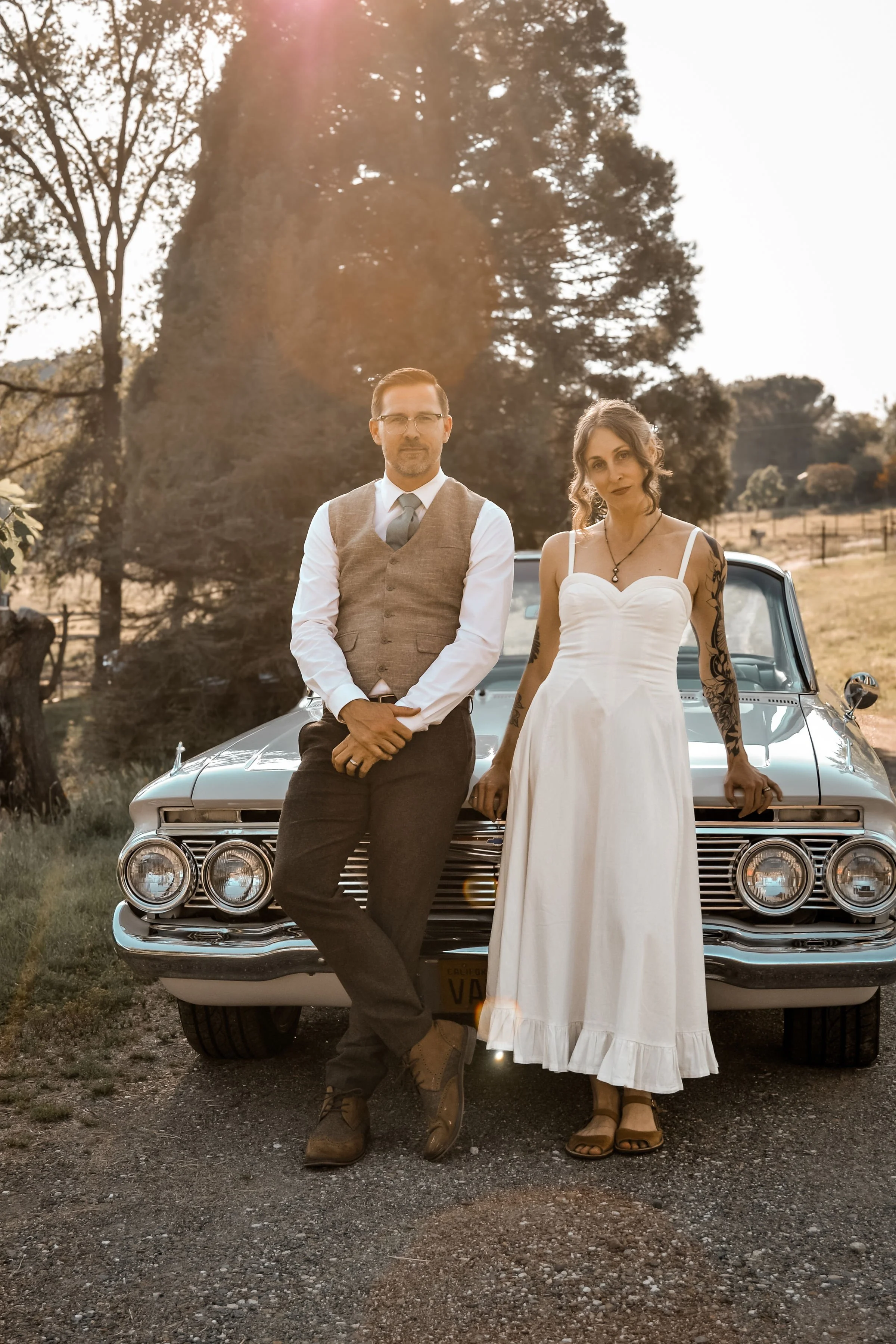 A man and a woman standing in front of a vintage car outdoors, with trees in the background during sunset.