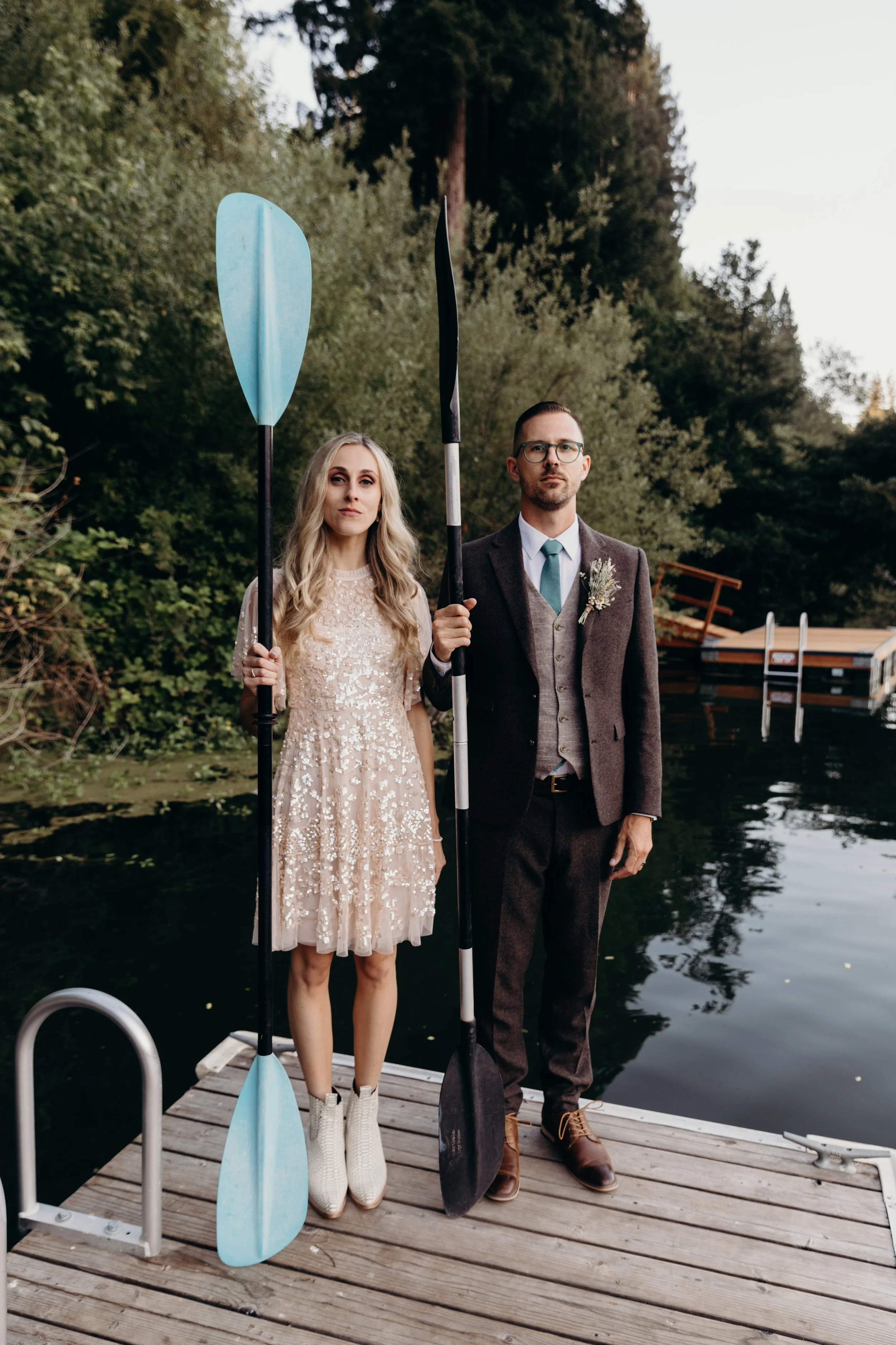 A couple standing on a dock holding paddles by a lake, surrounded by trees.