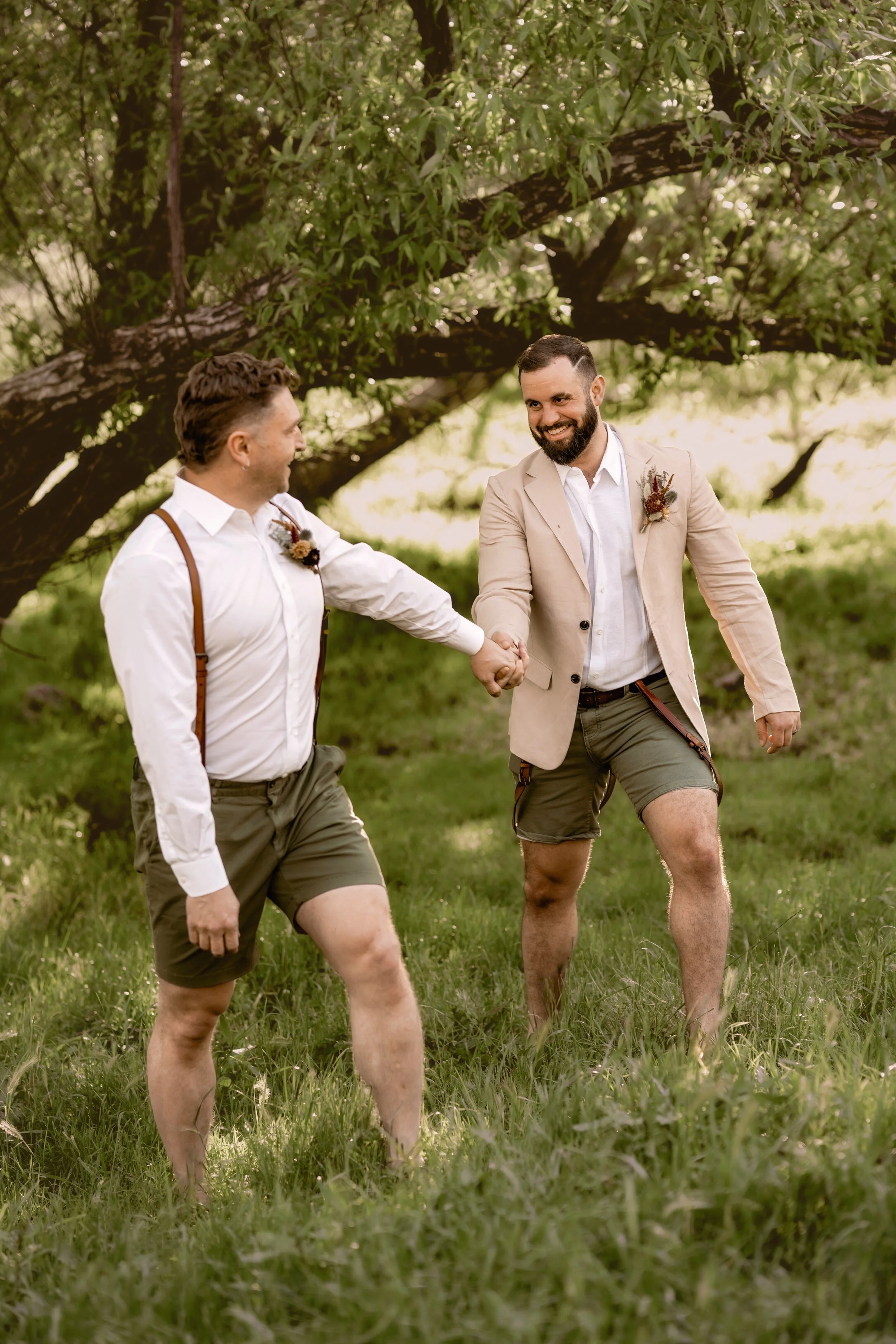 Two men are smiling and holding hands outdoors in a grassy field with trees in the background, dressed in semi-formal attire with suspenders and boutonnières.