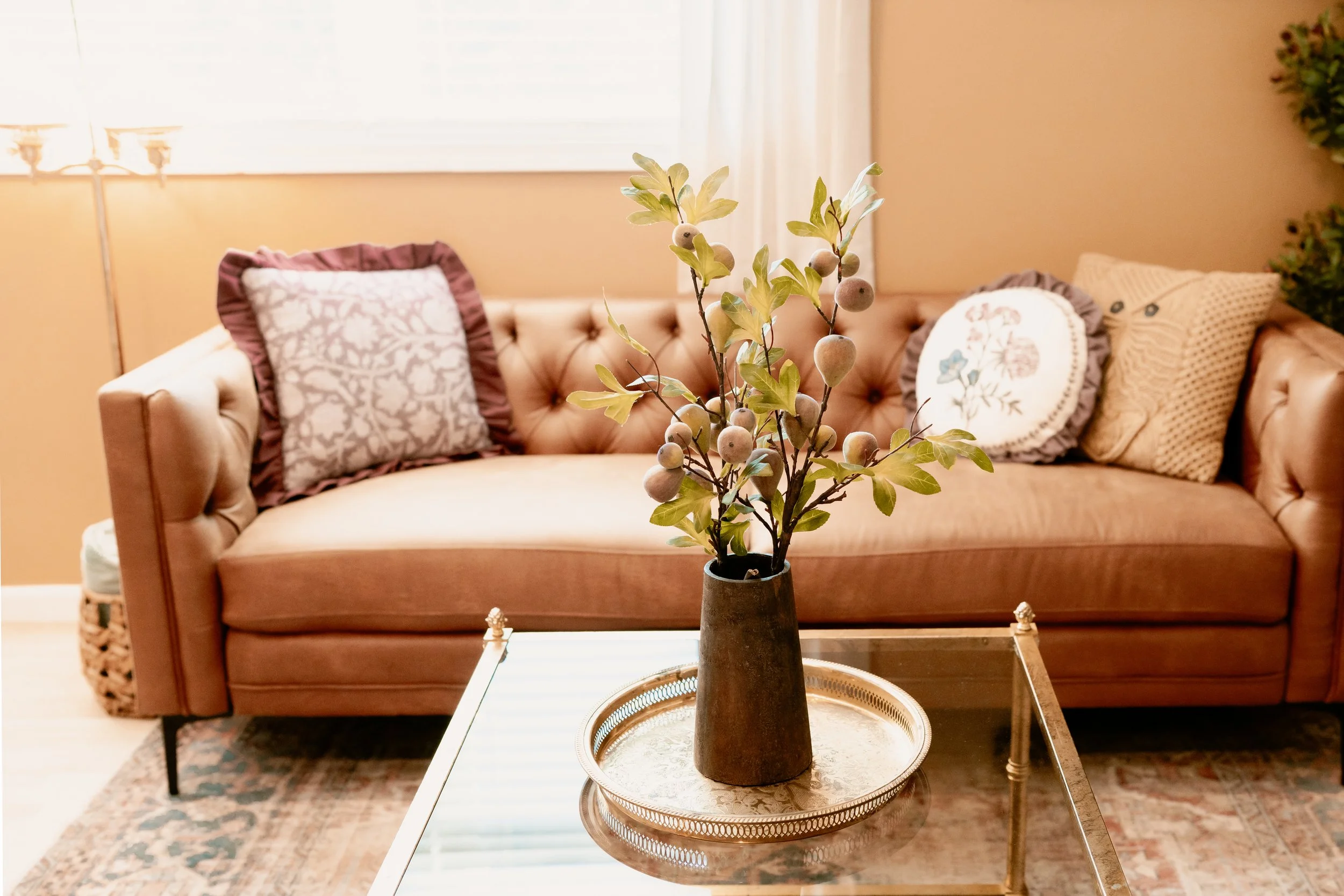 A living room with a beige tufted sofa, decorative pillows, a glass coffee table with a tall vase of greenery, a patterned rug, and sunlight streaming through a window.